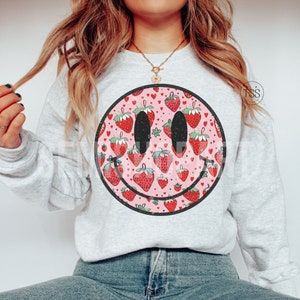 May include: A light gray sweatshirt with a pink and black smiley face design. The smiley face is filled with a pattern of red strawberries and pink hearts.