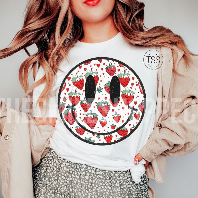 May include: White t-shirt with a black and white smiley face graphic. The smiley face is filled with a pattern of red strawberries and white hearts.