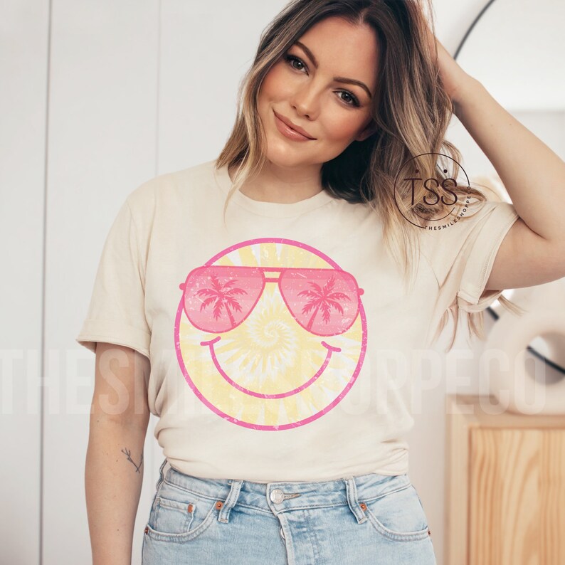 May include: A beige t-shirt with a pink and yellow smiley face graphic. The smiley face is wearing pink sunglasses with palm trees reflected in the lenses. The text "TSS The Smile Shop" is visible on the back of the shirt.