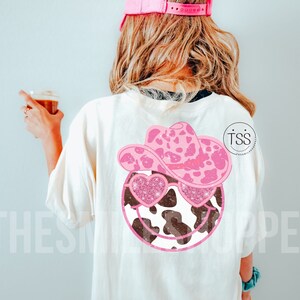 May include: White t-shirt with a pink cow print smiley face wearing a pink cow print hat and pink glitter heart sunglasses. The text "TSS" is on the right side of the shirt.