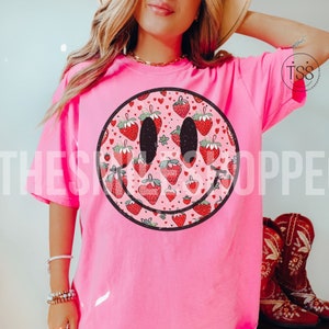 May include: Pink t-shirt with a black smiley face graphic. The smiley face is filled with a pattern of red strawberries and pink hearts.