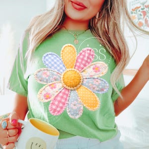 May include: A light green t-shirt featuring a large, colorful daisy applique. The daisy has petals in various patterns, including gingham and floral prints, with a yellow center. The shirt is a casual style.
