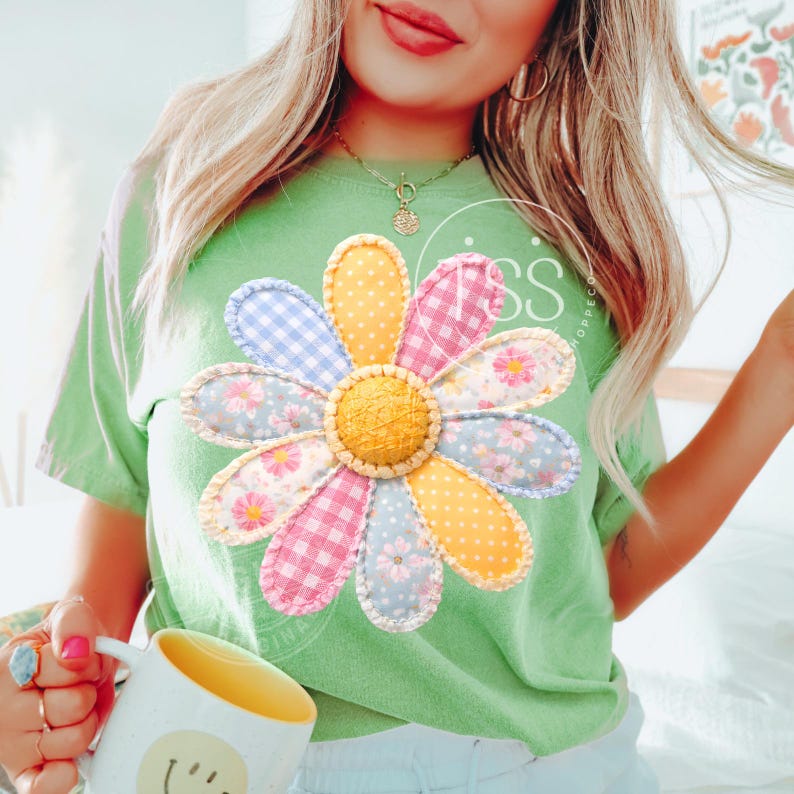 May include: A light green t-shirt featuring a large, colorful daisy applique. The daisy has petals in various patterns, including gingham and floral prints, with a yellow center. The shirt is a casual style.