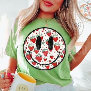 May include: Green t-shirt with a black and white smiley face design. The smiley face is filled with a pattern of red strawberries and white hearts.
