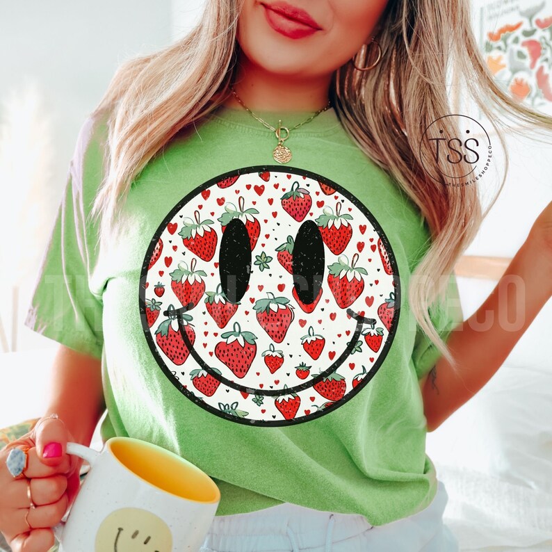 May include: Green t-shirt with a black and white smiley face design. The smiley face is filled with a pattern of red strawberries and white hearts.