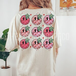 May include: A beige t-shirt with a pattern of nine pink and red smiley faces with green leaves. Each smiley face is filled with a pattern of small red hearts and strawberries.