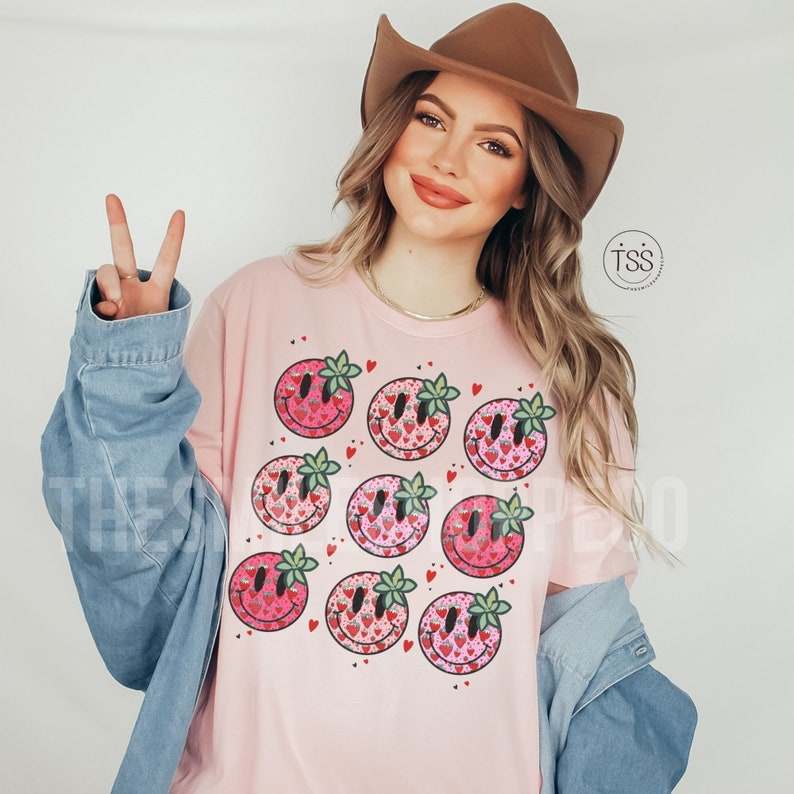 May include: Pink t-shirt with a pattern of smiling faces made of strawberries. Each face has a green leaf on top. The shirt is worn by a person with long brown hair and a brown cowboy hat.