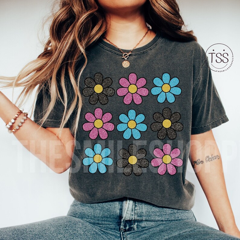 May include: A gray t-shirt with a pattern of nine glittery daisy flowers in black, pink, and blue. The flowers are arranged in a 3x3 grid.
