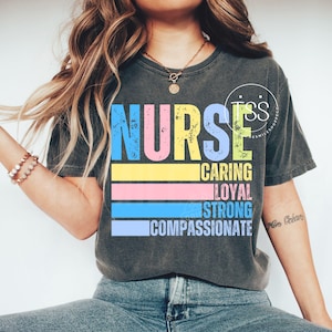 Op de afbeelding: Donkergrijs t-shirt met het woord "NURSE" in grote, kleurrijke letters. Daaronder staan de woorden "CARING, LOYAL, STRONG, COMPASSIONATE" op horizontale strepen. Het shirt heeft een vintage, distressed look.