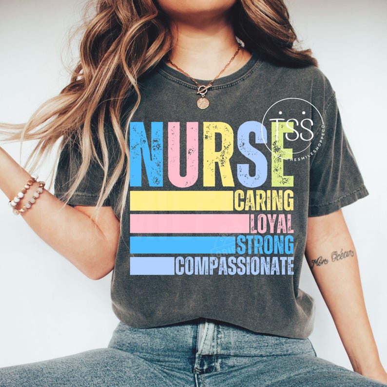 Op de afbeelding: Donkergrijs t-shirt met het woord "NURSE" in grote, kleurrijke letters. Daaronder staan de woorden "CARING, LOYAL, STRONG, COMPASSIONATE" op horizontale strepen. Het shirt heeft een vintage, distressed look.