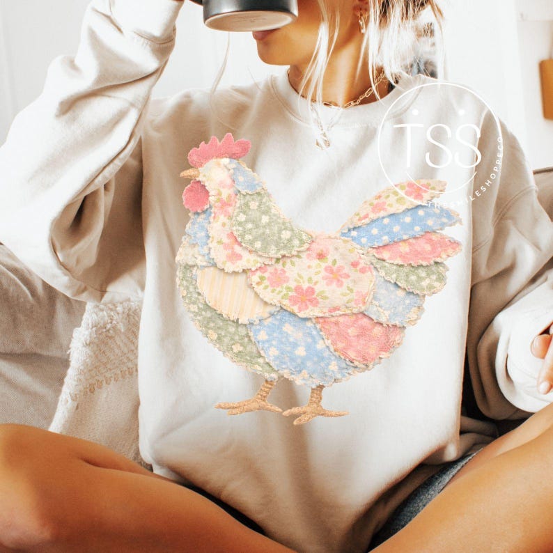 May include: A cream-colored sweatshirt featuring a colorful patchwork chicken design. The chicken has a red comb and wattle, with wings and body made of various pastel-colored fabric patches. The design is centered on the front of the sweatshirt.
