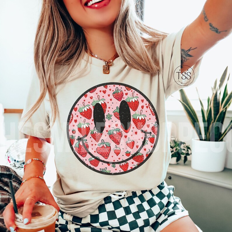 May include: A white t-shirt with a pink and red smiley face design. The smiley face is filled with a pattern of strawberries and hearts.