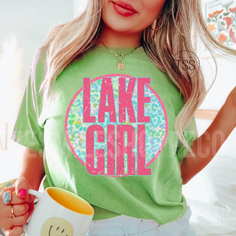 May include: A green t-shirt with a pink circle graphic that says "LAKE GIRL" in a distressed font. The circle graphic has a blue, green, and yellow leopard print pattern.