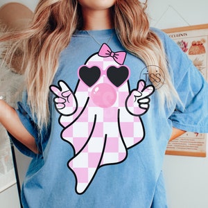 K&ouml;nnte beinhalten: Ein blaues T-Shirt mit einem rosa-wei&szlig; karierten Geist, der eine Sonnenbrille und eine rosa Schleife tr&auml;gt. Der Geist bl&auml;st eine rosa Blase.