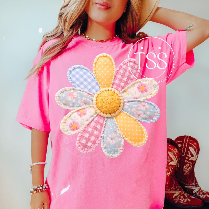 May include: A bright pink t-shirt featuring a large, colorful daisy applique. The daisy has petals in various patterns and colors, including blue gingham, yellow polka dots, and floral prints. The shirt is a comfortable, oversized fit.