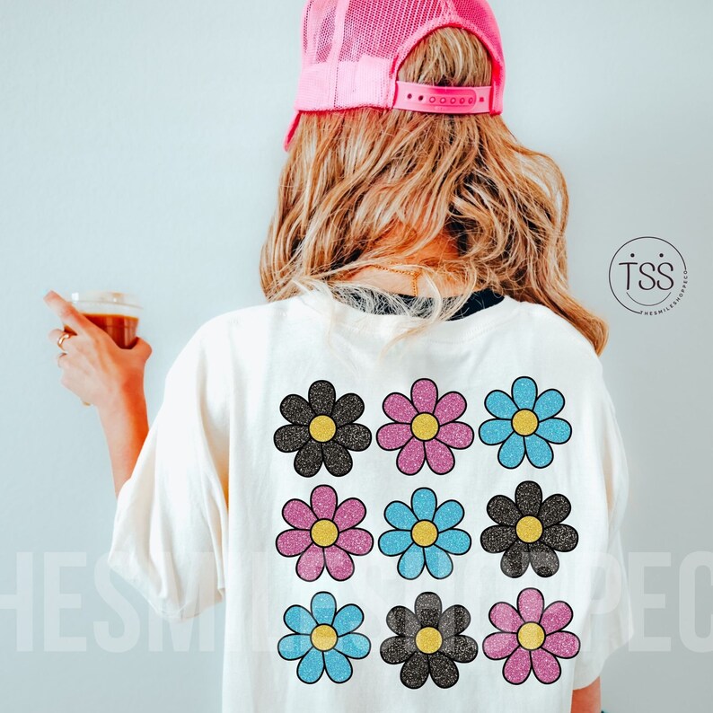 May include: A white t-shirt with a design of nine glittery daisies in black, pink, and blue colors.