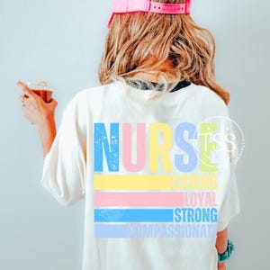 Op de afbeelding: Cr&egrave;mekleurig T-shirt met het woord "NURSE" in grote, kleurrijke letters op de rug. Onder het woord "NURSE" staan de woorden "CARING", "LOYAL", "STRONG" en "COMPASSIONATE" in een kleurrijk horizontaal streepontwerp.