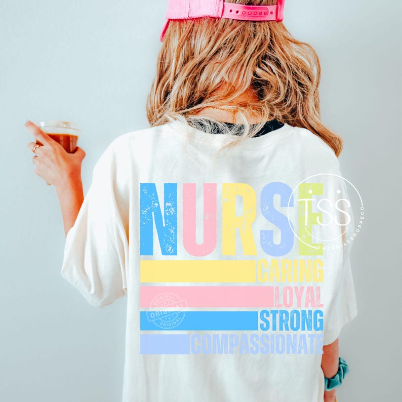 Op de afbeelding: Cr&egrave;mekleurig T-shirt met het woord "NURSE" in grote, kleurrijke letters op de rug. Onder het woord "NURSE" staan de woorden "CARING", "LOYAL", "STRONG" en "COMPASSIONATE" in een kleurrijk horizontaal streepontwerp.