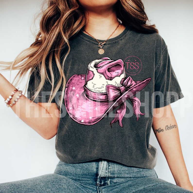 May include: A grey t-shirt featuring a pink and white cow print cowboy hat with a pink bow. The hat has white stars on it. The shirt has the text "TSS" in pink.