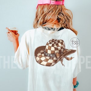 Pu&ograve; includere: Una maglietta bianca con un grafico di cappello da cowboy a scacchi marroni e bianchi sul retro. Il cappello ha una fascia marrone con una fibbia argentata e un nastro marrone legato attorno alla corona.