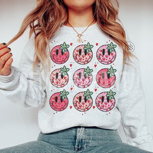 May include: A gray sweatshirt with a pattern of nine smiley faces. Each smiley face is filled with a pink and red strawberry pattern.  The smiley faces have green leaves on top.  There are small red hearts scattered throughout the pattern.