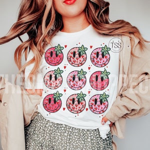 May include: White t-shirt with a pattern of nine red and pink smiley faces with green leaves. Each smiley face is filled with a pattern of small red hearts and strawberries.