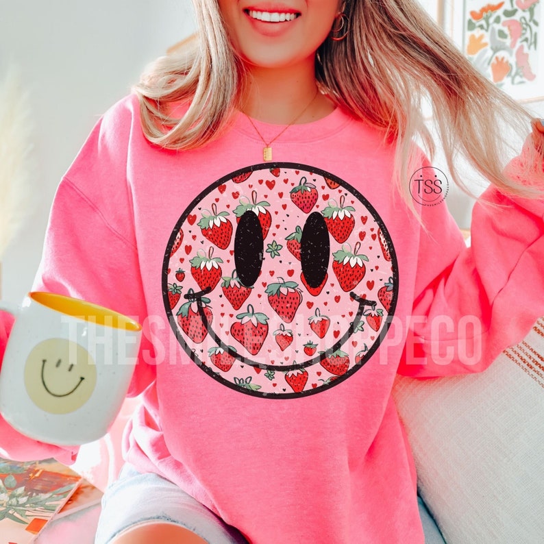 May include: Pink sweatshirt with a black and white smiley face design. The smiley face is filled with a pattern of red strawberries and white hearts.