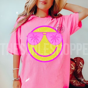 May include: A bright pink t-shirt featuring a yellow smiley face wearing pink sunglasses. The sunglasses reflect palm trees. The text "THE SMILES SHOPPE" is visible on the shirt. The shirt is a comfortable, casual style.