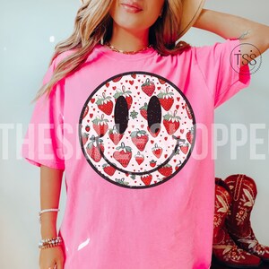 May include: Pink t-shirt with a large smiley face graphic. The smiley face is filled with a pattern of red strawberries and white hearts.