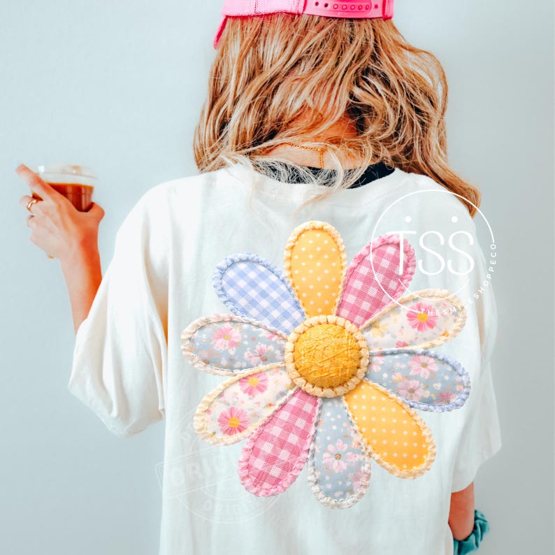 May include: White t-shirt with a large, colorful daisy applique on the back. The daisy features petals in various patterns and colors, including pink gingham, blue gingham, and floral prints. The shirt is worn by a person with blonde hair and a pink cap.