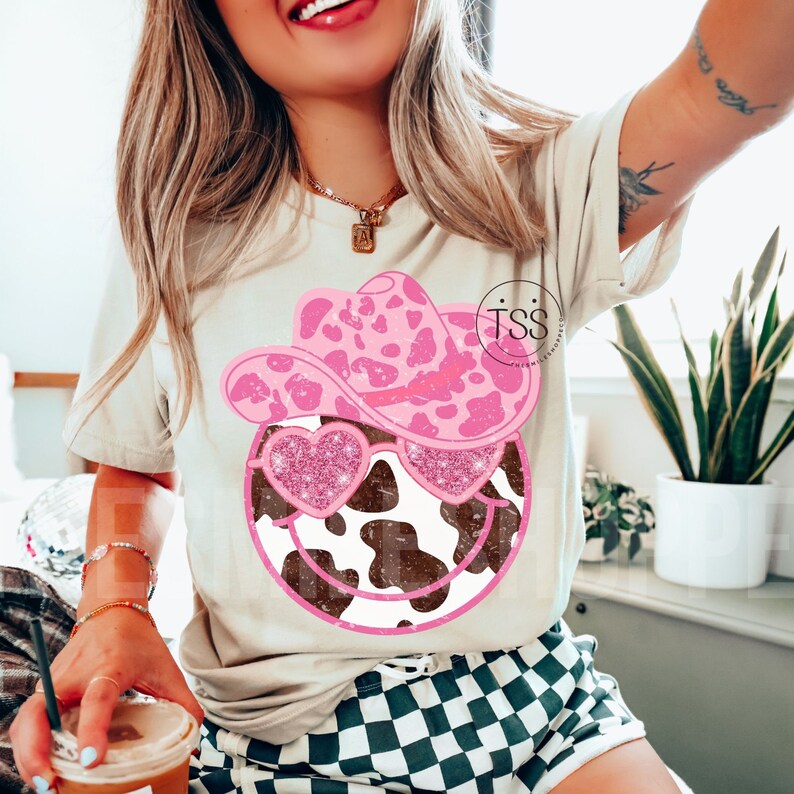May include: A white t-shirt with a pink and brown cow print smiley face wearing a pink cowboy hat and pink glitter sunglasses. The text "TSS" is visible on the shirt.