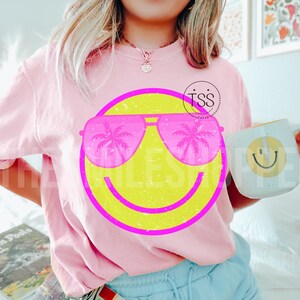 May include: Pink crew neck sweatshirt featuring a large yellow smiley face with pink sunglasses reflecting palm trees. The sweatshirt is paired with a white mug with a yellow smiley face.