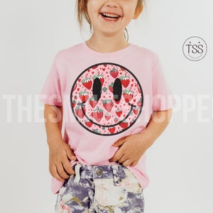 May include: A pink t-shirt with a smiley face design. The smiley face is filled with a pattern of red strawberries. The t-shirt is worn by a child.