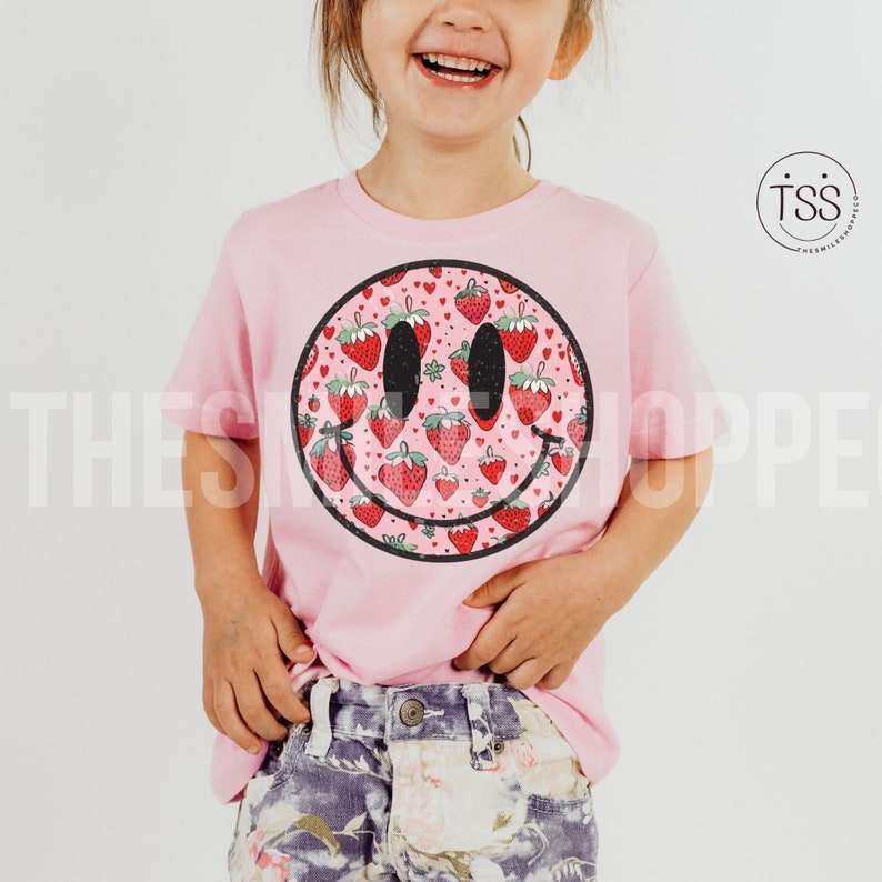 May include: A pink t-shirt with a smiley face design. The smiley face is filled with a pattern of red strawberries. The t-shirt is worn by a child.