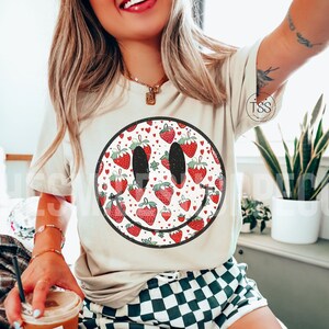 May include: A white t-shirt with a black smiley face graphic. The smiley face is filled with a pattern of red strawberries and small red hearts.