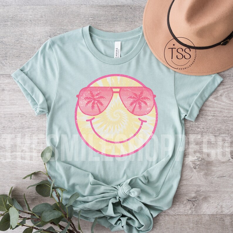 May include: A light blue t-shirt with a yellow smiley face wearing pink sunglasses with palm trees reflected in the lenses. The smiley face has a tie-dye pattern.