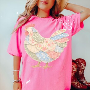 May include: A bright pink t-shirt featuring a colorful patchwork chicken design. The chicken has a red comb and wattle, with wings made of floral and polka dot patterned fabric. The shirt is a comfortable, oversized fit.