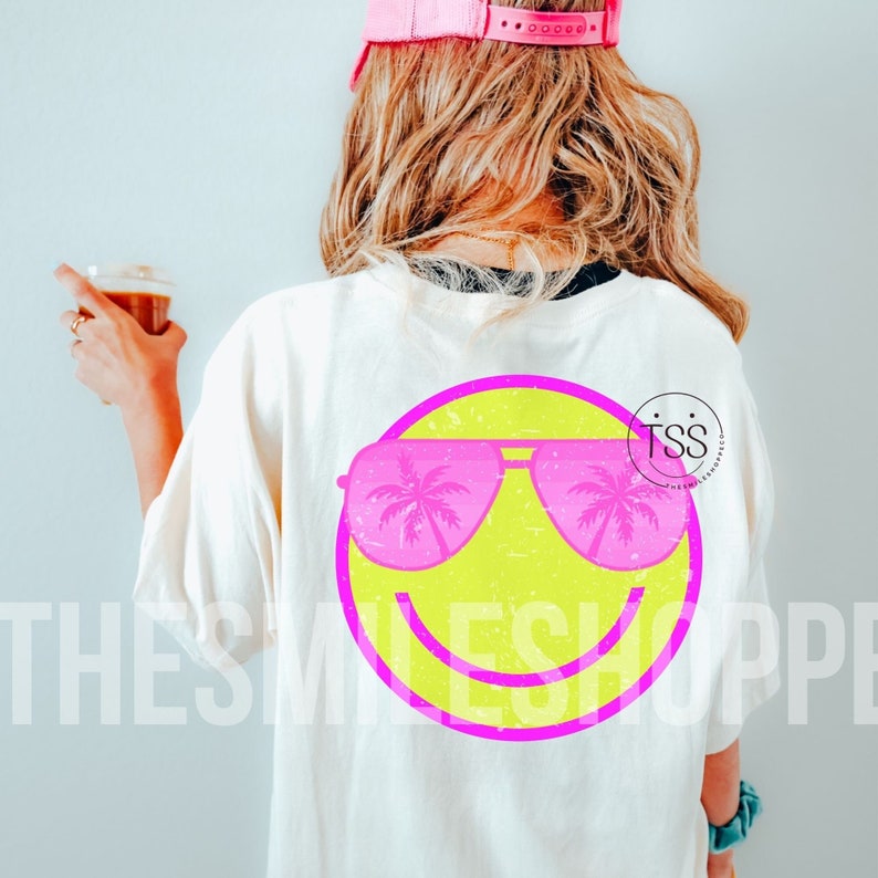 May include: Cream-colored t-shirt featuring a neon yellow smiley face with pink sunglasses reflecting palm trees. The design is inside a pink circle. The text "THE SMILE SHOPPE" is printed on the shirt.