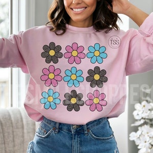 May include: A pink sweatshirt with a design of nine glitter daisies in black, pink, and blue. The daisies are arranged in three rows of three. The sweatshirt has the text "TSS" in a circle on the right side of the chest.