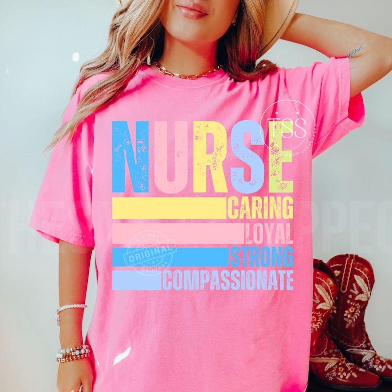 Op de afbeelding: Een felroze t-shirt met het woord "NURSE" in blauwe, gele en groene letters. Daaronder staan de woorden "CARING", "LOYAL", "STRONG" en "COMPASSIONATE". Het shirt heeft een relaxte pasvorm en is gemaakt van zacht materiaal.