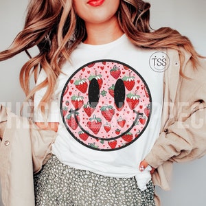 May include: White t-shirt with a pink and red smiley face graphic. The smiley face is filled with a pattern of strawberries and hearts.