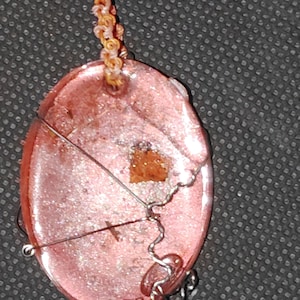May include: An oval-shaped pendant with a shimmering pink, resin-like surface containing a small embedded brown fragment. The pendant is wrapped in thin silver-colored wire and hangs from a braided cord featuring light blue and orange threads.