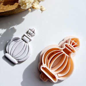 Op de afbeelding: Een licht perzikkleurige plastic koekjesvorm in de vorm van een vintage parfumflesje met een gefacetteerde dop, vergezeld van een kleine, lichtgrijze klei-uitsparing van hetzelfde parfumflesontwerp, met de reliëfdetails zichtbaar.