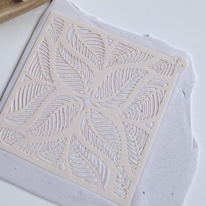 May include: A light beige stencil with a symmetrical leaf design. The stencil is placed on a light blue surface. The leaf pattern is repeated in a square arrangement, with each leaf having detailed vein lines. The stencil is likely used for crafts or art projects.