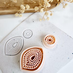 Op de afbeelding: Een verzameling van drie lichtroze kleisnijders met ingewikkelde bloemen- en bladerontwerpen. De snijders liggen op een gespikkeld wit oppervlak, met gedroogde bloemen en een houten plank op de achtergrond. Geschikt voor knutselen of bakken.