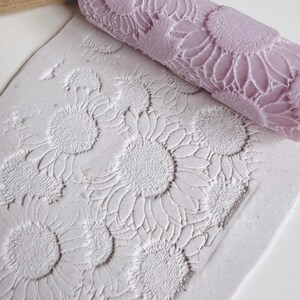 May include: A lilac rolling pin with a sunflower pattern sits beside a sheet of clay with the same embossed design. The rolling pin is cylindrical and features a repeating sunflower motif. The clay sheet displays the sunflower pattern in relief.