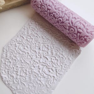 May include: A lavender rolling pin with an embossed floral design rests beside a white clay tile with a matching embossed floral pattern. The rolling pin is cylindrical, and the tile is irregularly shaped. The items are on a white surface.