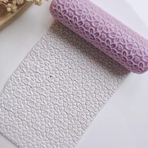 Peut inclure: Un rouleau à pâtisserie lavande avec un motif floral en forme de cœur repose sur une pièce d'argile avec le même motif en relief. L'argile est blanche et le rouleau est de couleur unie. L'image est un gros plan.