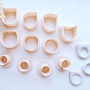 May include: A selection of light pink polymer clay cutters in diverse shapes, such as rounded rectangles, circles, and teardrops. Some cutters feature millimetre measurement markings. The cutters are arranged on a white surface, with dried flowers in the top left corner.