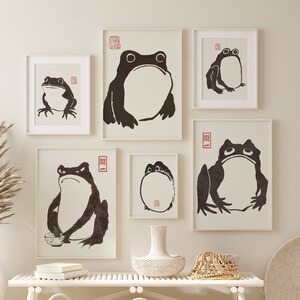 May include: A white wooden bench with two white wicker baskets underneath. There are five framed black and white prints of frogs on the wall above the bench. The prints are arranged in a symmetrical pattern. A vase with dried pampas grass sits on the floor to the left of the bench.